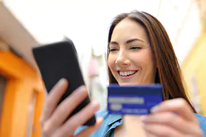 Smiling woman outdoors holds a smartphone in one hand and a blue credit card in the other, as if making a payment.